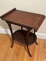 Full view of vintage side table showing leather top, scroll edge and carved legs with lower wooden shelf.