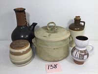 Five vintage stoneware pottery items displayed on a white surface including a large lidded pot, leather-wrapped pitcher, brown jug, patterned mug, and small pot