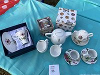 All five tea and coffee sets displayed on a blue cloth: floral tea cup/saucer boxed, coffee mug with lid boxed, white teapot and cup, two espresso cups with shells, seashell stacking set boxed.
