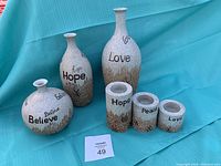 Full set of six ceramic items on blue background: three vases with narrow mouths labeled "Love," "Hope," and "Believe," and three cylindrical tealight candle holders labeled "Hope," "Peace," and "Love."
