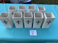 Nine white rectangular ceramic vases with silver crosses, arranged on a blue tablecloth, showing full set.