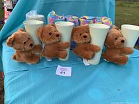 Eight light green FTD brand vases each paired with brown teddy bears hugging them, displayed on blue fabric.