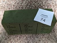 Single green Oasis floral foam brick showing top and side with pre-cut grooves for easy breaking, placed on carpet background with auction tag on top.