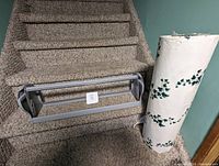 Photo showing a gray wall mounted paper cutter without dowel and a large roll of white paper with English Ivy leaf pattern on it, both placed on carpeted stairs.