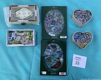 All six stained glass items displayed on a blue fabric background: two rectangular keepsake boxes, two heart-shaped keepsake boxes, and two oval sun catchers in original packaging.