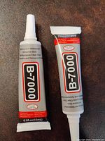Two B-7000 jeweler's glue tubes side by side on a brown surface, showing the full tube and labeling.