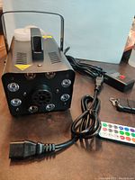 Front view of black fog machine with eight surrounding LED lights, power cord, remote control, and LED light controller on a dark surface.