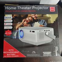 Box displaying RCA home theater projector branding, showing features including Bluetooth, up to 150" picture size, 2 HDMI inputs, HD 720p resolution, and rechargeable battery.