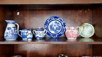 Photo showing Blue Willow transferware pitcher, salt shaker, cream and sugar, red transferware Enoch Wood pitcher, Nino-Po china plate, and Bavarian nappy dish on a shelf.