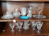 Photo shows the full vintage crystal stemware and serving ware set arranged on a shelf, including the goblets, pitcher, domes, platter, and blue dish.