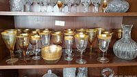 Wide shot showing entire collection of gold rimmed stemware glasses, bowls, and crystal decanter arranged on shelf.