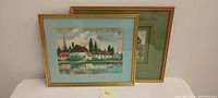Two vintage watercolor paintings framed in gold-colored frames on a white surface. The paintings depict lakeside houses and natural landscapes.