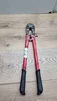 Front view of red bolt cutters with black rubber grips, showing the tool laid flat on wood surface.