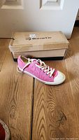 Pink Burnetie sneaker placed on wooden floor next to its original box, showing side and top view of the shoe.
