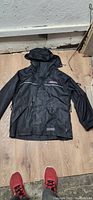 Front view of Dakota black winter jacket laid flat on floor showing front zipper, snap-button storm flap, hood with drawstrings, and two zippered front pockets.