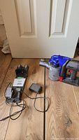 Dyson handheld vacuum with wall mount charger and charging adapter on wooden floor near white door.