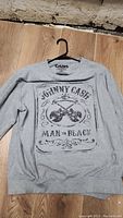 Gray Johnny Cash sweater laid flat on a wooden floor showing the front design with crossed guitars and text