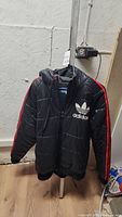 Front view of black Adidas winter coat with hood, red sleeve stripes, and white Adidas logo on chest.