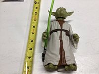 Yoda figure shown next to a measuring tape indicating approximately 10 inches tall. Figure is green with white Jedi robe and is holding a green lightsaber.