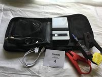 Photo of a portable battery booster device, including a silver and black rectangular unit with cables: car cigarette lighter cable, large red and black clamps, and a white USB cable, all inside a black carrying case.