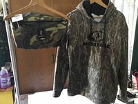 Photo of Mossy Oak camouflage pullover hoodie and matching camouflage bag hanging on wooden hangers.