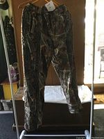 Front view of the camouflage pants hanging on a hanger, showing natural woodland pattern with brown, green, and gray tones, placed in an indoor setting with mirror and furniture visible.