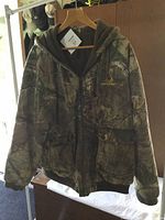 Front view of camo lined coat hanging on a hanger showing zip front, pockets, and hood