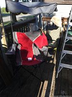 Red and gray foldable camping chair with canopy and cup holder taken in sunlight on a wooden porch.