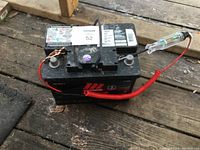 Motomaster car battery with red coiled wire attached to positive terminal and tester tool on right terminal. Shown on wooden surface with labels visible.