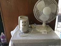 Photo of a portable electric heater and a white oscillating table fan placed on a table.