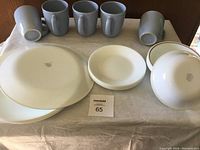 Overall view of 3 Corelle cups, 1 plate, 1 bowl, and 1 large plate, exhibiting mixed white dishware with visible wear marks, showing tops and bottoms of items.