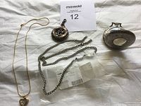 Photo of lot showing five jewelry pieces: three pendants including a gold-tone heart pendant with a clear gem, two silver chains, and an ornate silver pocket watch.