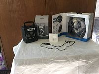 Three items on white fabric: boxed SoundLogic XT over-ear headphones, portable Bluetooth speaker with handle and control panel, wired earbuds on table.