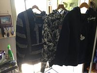 Three mens pullovers hanging on wooden hangers along a bright window showing stripes, floral pattern, and Mickey Mouse embroidery.