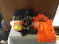 The photo shows a collection of safety wear items including a high-visibility orange shirt, two camouflage Hard Hat covers, elastic safety bands, and a face covering.