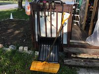 Front view showing both the yellow plow style shovel with wooden handle leaning against a large black push shovel with metal handle near an outdoor wooden structure.