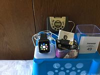 Photo showing Bluetooth smart watch on charging dock with USB cable, Proscan smart watch box, and Panasonic portable radio cassette player in background.