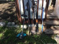 Photo showing set of yard tools lined up against a shed including electric weed wacker, crowbar, shovel, garden fork, and weed puller.