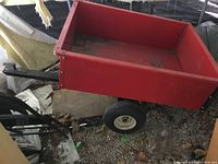 Red metal garden tractor dump trailer with black tow handle and two wheels, showing side and top views with dirt and signs of use inside the bed.