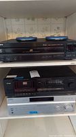 Sony 5 CD player on top shelf, Teac cassette player in the middle shelf, and Sony receiver on bottom shelf all stacked within a cubby with floral wallpaper background. Two blue handheld devices on top of the Sony CD player.