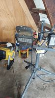 Photo showing yellow Stanley portable heater on floor, black front grill and handle visible, next to metal tripod stand with attached work light and some paint cans under stand.