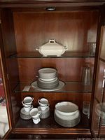 Glass cabinet with Noritake Crestwood Platinum dinnerware set including stacked dinner, salad, and dessert plates, cups with saucers, bowls, plus one large Wedgwood white covered serving dish on top shelf.
