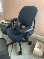 Front view of blue Herman Miller Leap office chair with adjustable armrests and small black personal table fan on the seat, next to a black metal mesh trash can on the floor.