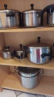 Multiple stainless steel pots and pans of different sizes with lids and black handles on wooden shelf.