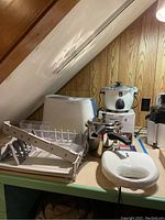 Photo showing the pressure cooker, white electric griddle, plastic trays, and a white rice cooker on a wooden shelf under stairs.