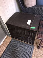 Front view of the rectangular dark brown plastic storage bin with closed hinged lid and rattan pattern sides.