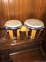 Pair of wooden bongo drums with natural skin heads and metal tuning rims on wooden surface, connected by a wooden block.