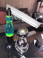 Three desk lamps on a glass table near a sofa: green-blue lava lamp, silver spherical moon lamp, beige fluorescent desk lamp.