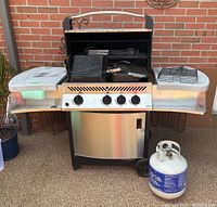 Napoleon stainless steel propane grill with side shelves, lid open, showing accessories in main chamber and propane cylinder beside.