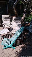 Two white folding plastic patio chairs with off-white cushions, green plastic folding lounge chair, white plastic side table, and green plastic chair clustered outside on a paved surface.
